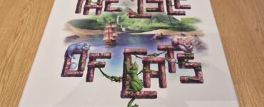 Board game box for "The Isle of Cats" featuring colorful illustrations of cats and a ship surrounded by a lush landscape, placed on a wooden table.