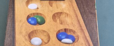 Mancala board game box featuring a solid wood game board, designed for 2 players aged 6 and up, including 48 colored stones and instructions.