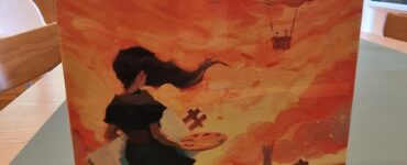 Box cover of the board game "Canvas," featuring a girl holding a paint palette against a vibrant orange sunset with whimsical clouds and a fantasy landscape in the background.