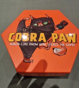 Hexagonal board game box for "Cobra Paw," featuring a cartoon ninja cat with green eyes, promoting a fun and competitive gameplay experience.