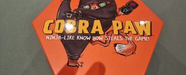 Hexagonal board game box for "Cobra Paw," featuring a cartoon ninja cat with green eyes, promoting a fun and competitive gameplay experience.