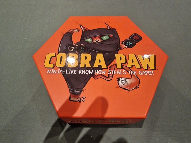 Hexagonal board game box for "Cobra Paw," featuring a cartoon ninja cat with green eyes, promoting a fun and competitive gameplay experience.