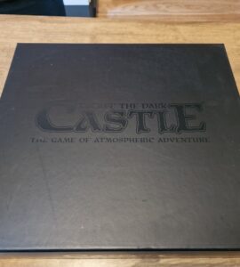 Black game box for "Escape the Dark Castle," featuring the title and tagline "The Game of Atmospheric Adventure" on the lid, placed on a wooden surface.