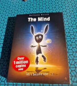 Box cover of the board game "The Mind" by Wolfgang Warsch, featuring a cartoon character with long ears and a playful expression. The design highlights the text "Over 1 million copies sold" in a red circle, promoting its popularity. The background has a glowing effect, emphasizing the game's engaging theme.