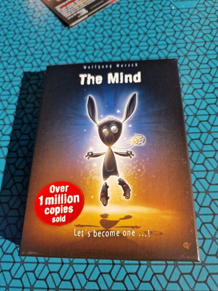 Box cover of the board game "The Mind" by Wolfgang Warsch, featuring a cartoon character with long ears and a playful expression. The design highlights the text "Over 1 million copies sold" in a red circle, promoting its popularity. The background has a glowing effect, emphasizing the game's engaging theme.