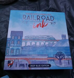 Cover of the board game "Railroad Ink: Deep Blue Edition" by Hjalmar Hach and Lorenzo Silva, featuring a whimsical illustration of a train crossing a bridge with buildings and trees in a blue color palette.