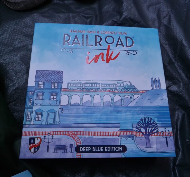 Cover of the board game "Railroad Ink: Deep Blue Edition" by Hjalmar Hach and Lorenzo Silva, featuring a whimsical illustration of a train crossing a bridge with buildings and trees in a blue color palette.