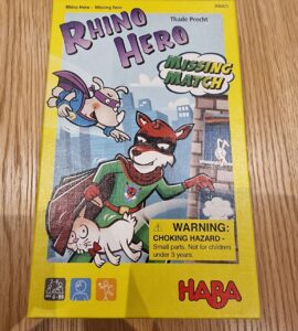 Colorful board game box for "Rhino Hero: Missing Match" by HABA, featuring cartoon characters including a superhero rhino, a cat, and a mouse. The box highlights the game's age recommendation (4-99 years) and includes a choking hazard warning. Ideal for family game nights and children's entertainment.