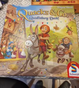 Colorful board game box for "Quacks & Co. Quedlinburg Dash!" by Wolfgang Warsch, featuring cheerful characters, a donkey, and festive decorations. The game is published by Schmidt.