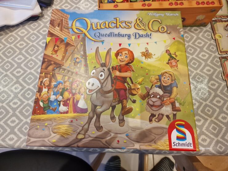 Colorful board game box for "Quacks & Co. Quedlinburg Dash!" by Wolfgang Warsch, featuring cheerful characters, a donkey, and festive decorations. The game is published by Schmidt.