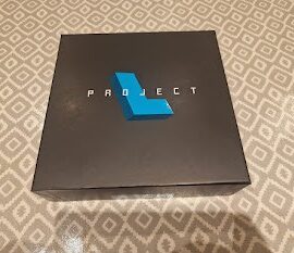 Black box with the word "PROJECT" in bold white letters and a blue geometric shape on top, placed on a patterned surface.