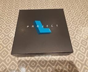 Black box with the word "PROJECT" in bold white letters and a blue geometric shape on top, placed on a patterned surface.