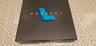 Black box with the word "PROJECT" in bold white letters and a blue geometric shape on top, placed on a patterned surface.