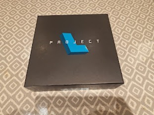 Black box with the word "PROJECT" in bold white letters and a blue geometric shape on top, placed on a patterned surface.