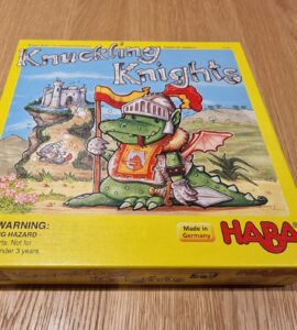 Colorful board game box for "Knuckling Knights" by HABA, featuring a cartoon dragon in knight armor, set against a whimsical castle backdrop. The box includes a warning label about choking hazards for small parts. Made in Germany.