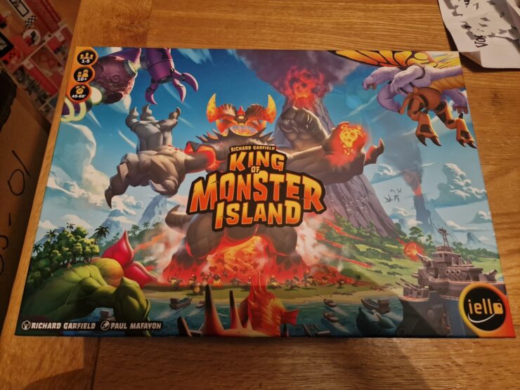 Box cover of the board game "King of Monster Island" featuring colorful artwork of giant monsters, a volcano, and a coastal landscape. Designed by Richard Garfield and Paul Mafavon, suitable for 2-5 players aged 10 and up, with a playtime of 45-60 minutes.