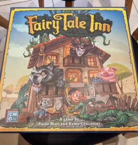 Colorful board game box for "Fairy Tale Inn" featuring whimsical characters like a wolf, a pig, and a wooden puppet, designed by Paolo Mori and Remo Conzadori. The illustration showcases a charming inn surrounded by fairy tale elements.