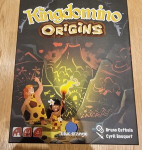 Box cover of Kingdomino Origins board game featuring vibrant artwork of two cave people, one holding a torch, set against a prehistoric backdrop. The game is designed for 2-4 players, ages 8 and up, with a playtime of approximately 25 minutes. Published by Blue Orange Games, created by Bruno Cathala and Cyril Bouquet.