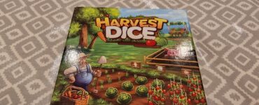 Box cover of the board game "Harvest Dice" designed by Danny Devine, featuring a farmer with a basket of vegetables, a lush garden with cabbage and tomatoes, and branding from Grey Fox Games.