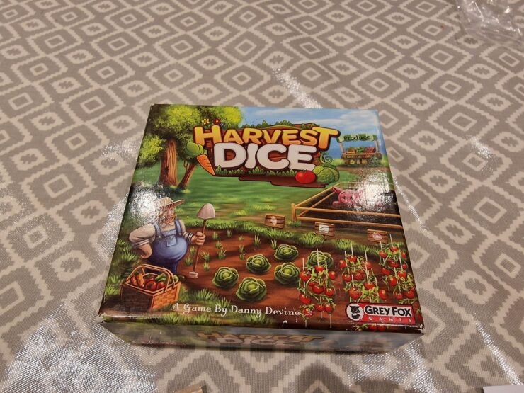 Box cover of the board game "Harvest Dice" designed by Danny Devine, featuring a farmer with a basket of vegetables, a lush garden with cabbage and tomatoes, and branding from Grey Fox Games.