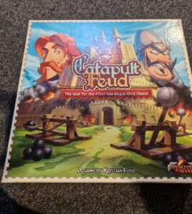 Catapult Feud board game box featuring colorful artwork of two characters, a castle, and catapults, designed by Kristian Fosh and published by Vesuvius Media. The tagline "The War for the Floor has Begun Once More!" highlights the game's competitive nature.