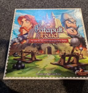 Catapult Feud board game box featuring colorful artwork of two characters, a castle, and catapults, designed by Kristian Fosh and published by Vesuvius Media. The tagline "The War for the Floor has Begun Once More!" highlights the game's competitive nature.