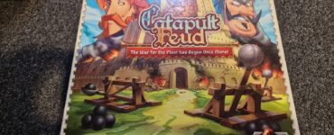 Catapult Feud board game box featuring colorful artwork of two characters, a castle, and catapults, designed by Kristian Fosh and published by Vesuvius Media. The tagline "The War for the Floor has Begun Once More!" highlights the game's competitive nature.