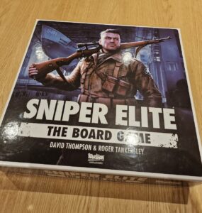 Box of Sniper Elite: The Board Game featuring artwork of a soldier with a rifle, designed by David Thompson and Roger Tankersley, placed on a wooden table.