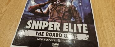Box of Sniper Elite: The Board Game featuring artwork of a soldier with a rifle, designed by David Thompson and Roger Tankersley, placed on a wooden table.