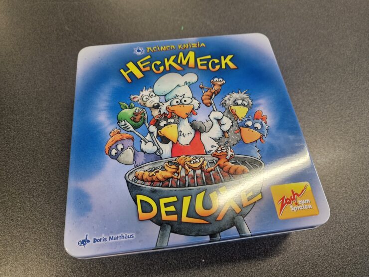 Board game box for "Heckmeck Deluxe" by Reiner Knizia, featuring cartoonish chickens and a chef grilling skewers, with vibrant colors and a playful design. Ideal for family game nights and gatherings.