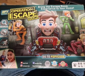 Colorful board game box for "Operation: Escape Room" featuring characters engaged in various escape activities. The design includes instructions to "Search, Solve, Escape" and highlights the game's social aspect for 2-6 players, ages 6 and up. The packaging also features social media handles and showcases awards, emphasizing the fun and interactive escape room experience at home.