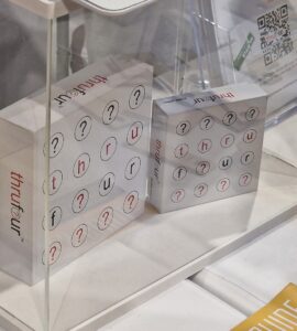 Two boxes of the game "thru four" displayed under a clear acrylic case, featuring a grid of letters and question marks. The game emphasizes word play and strategy, appealing to puzzle enthusiasts. Nearby, promotional material and a QR code are visible, suggesting additional information about the game.
