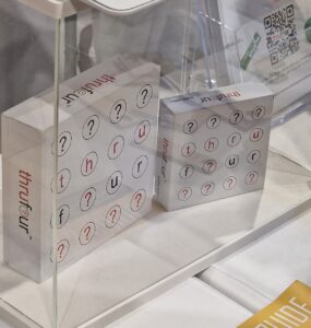 Two boxes of the game "thru four" displayed under a clear acrylic case, featuring a grid of letters and question marks. The game emphasizes word play and strategy, appealing to puzzle enthusiasts. Nearby, promotional material and a QR code are visible, suggesting additional information about the game.