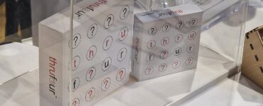 Two boxes of the game "thru four" displayed under a clear acrylic case, featuring a grid of letters and question marks. The game emphasizes word play and strategy, appealing to puzzle enthusiasts. Nearby, promotional material and a QR code are visible, suggesting additional information about the game.