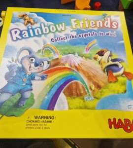 Colorful board game box titled "Rainbow Friends" by HABA, featuring a playful koala and a duck, with a vibrant rainbow and mountains in the background. The box emphasizes collecting crystals to win and includes a warning about small parts. Suitable for ages 4 and up.