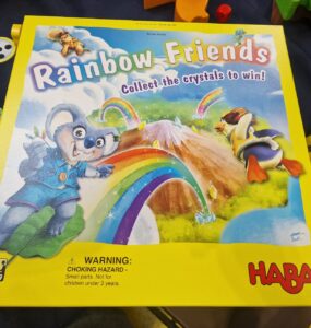 Colorful board game box titled "Rainbow Friends" by HABA, featuring a playful koala and a duck, with a vibrant rainbow and mountains in the background. The box emphasizes collecting crystals to win and includes a warning about small parts. Suitable for ages 4 and up.