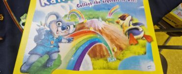 Colorful board game box titled "Rainbow Friends" by HABA, featuring a playful koala and a duck, with a vibrant rainbow and mountains in the background. The box emphasizes collecting crystals to win and includes a warning about small parts. Suitable for ages 4 and up.