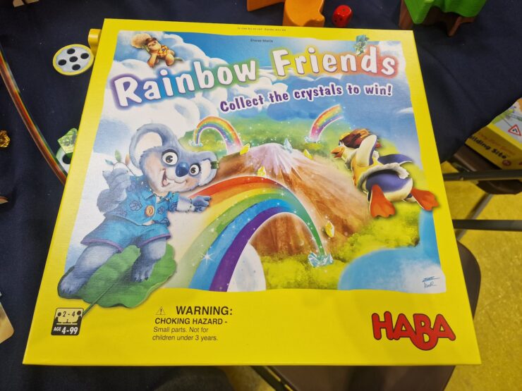 Colorful board game box titled "Rainbow Friends" by HABA, featuring a playful koala and a duck, with a vibrant rainbow and mountains in the background. The box emphasizes collecting crystals to win and includes a warning about small parts. Suitable for ages 4 and up.