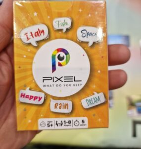 Image of the board game "Pixel," featuring a colorful box design with the title prominently displayed. The box includes playful words like "Italy," "Fish," "Space," "Happy," "Rain," and "Dream," indicating the game's creative theme. It is suitable for ages 5 and up, accommodating 1-8 players, and designed for quick play sessions of 5 minutes or more.