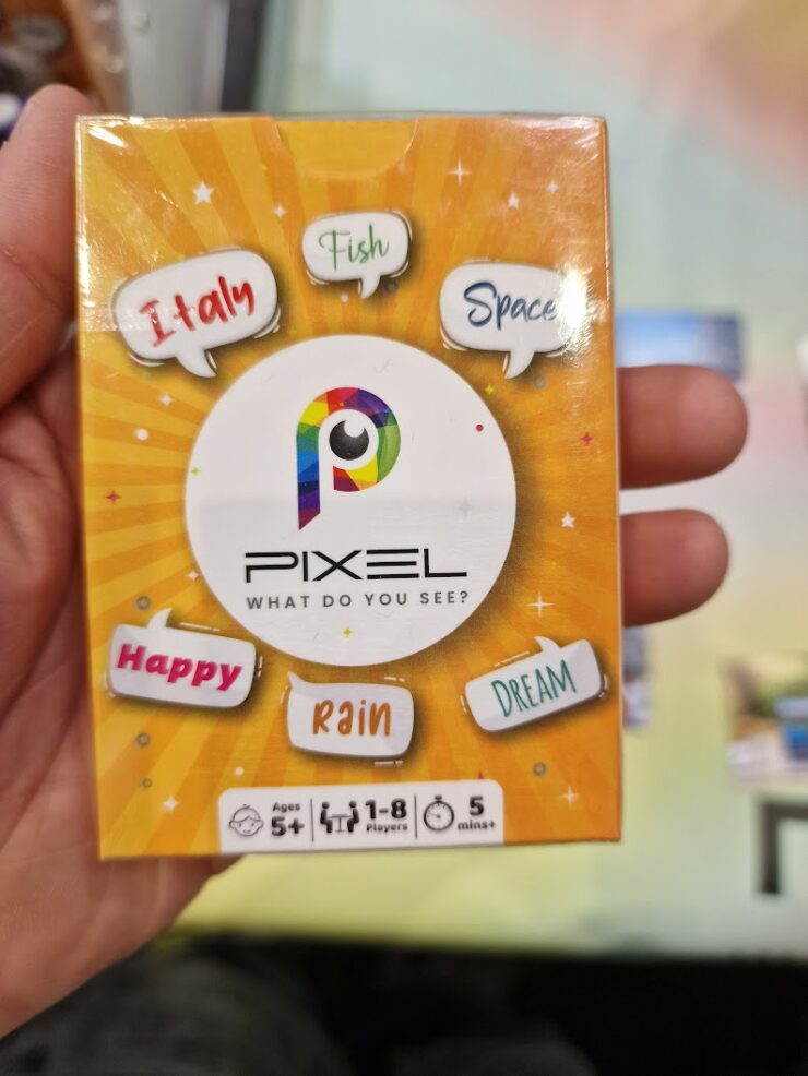 Image of the board game "Pixel," featuring a colorful box design with the title prominently displayed. The box includes playful words like "Italy," "Fish," "Space," "Happy," "Rain," and "Dream," indicating the game's creative theme. It is suitable for ages 5 and up, accommodating 1-8 players, and designed for quick play sessions of 5 minutes or more.