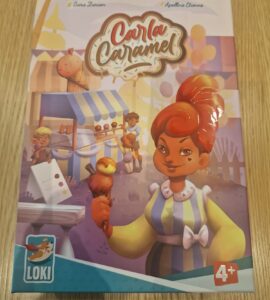 Colorful board game box for "Carla Caramel" featuring a character holding a caramel treat, designed for ages 4 and up. The background includes a festive market scene with children and food stalls.