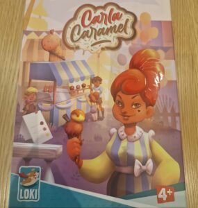 Colorful board game box for "Carla Caramel" featuring a character holding a caramel treat, designed for ages 4 and up. The background includes a festive market scene with children and food stalls.
