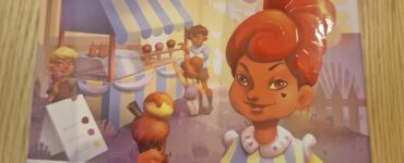 Colorful board game box for "Carla Caramel" featuring a character holding a caramel treat, designed for ages 4 and up. The background includes a festive market scene with children and food stalls.