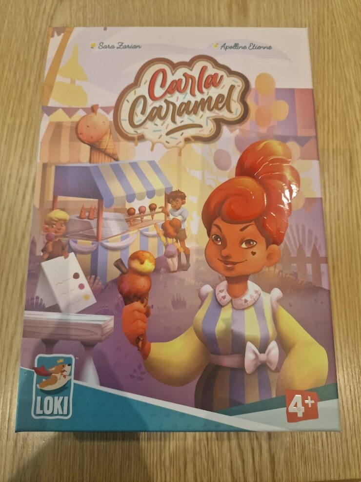 Colorful board game box for "Carla Caramel" featuring a character holding a caramel treat, designed for ages 4 and up. The background includes a festive market scene with children and food stalls.