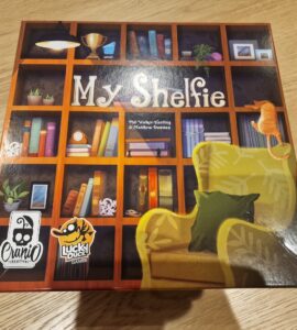 Board game box of "My Shelfie," featuring a colorful bookshelf design with various decorative items, a cozy green chair, and a playful cat, published by Cranio Creations and Lucky Duck Games.