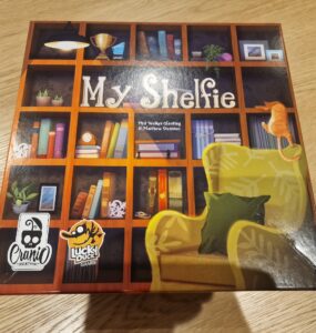 Board game box of "My Shelfie," featuring a colorful bookshelf design with various decorative items, a cozy green chair, and a playful cat, published by Cranio Creations and Lucky Duck Games.