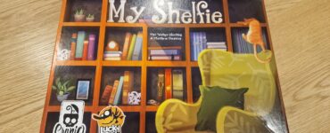 Board game box of "My Shelfie," featuring a colorful bookshelf design with various decorative items, a cozy green chair, and a playful cat, published by Cranio Creations and Lucky Duck Games.
