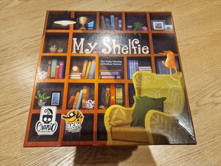 Board game box of "My Shelfie," featuring a colorful bookshelf design with various decorative items, a cozy green chair, and a playful cat, published by Cranio Creations and Lucky Duck Games.