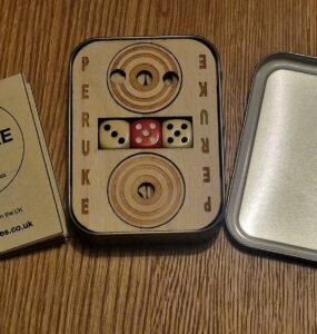 Peruke board game set displayed on a wooden surface, featuring a wooden game board with dice, a rules booklet, and a metal tin. Ideal for family game nights and strategic play, emphasizing handcrafted quality from the UK.