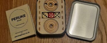 Peruke board game set displayed on a wooden surface, featuring a wooden game board with dice, a rules booklet, and a metal tin. Ideal for family game nights and strategic play, emphasizing handcrafted quality from the UK.